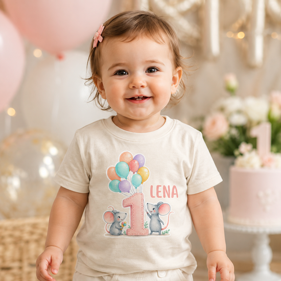 Lächelndes Kleinkind trägt ein personalisiertes cremefarbenes Geburtstagsshirt mit Name Lena, rosa Zahl 1, bunten Ballons und Maus Motiv. Ideales Shirt für den ersten Geburtstag eines Babys, Familienfeier oder Geburtstagsshooting im Boho Stil.