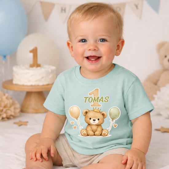 Baby trägt grünes personalisiertes T-Shirt mit Name Tomas und Zahl 1, süßer Bär mit Ballons, perfekte Outfitidee für ersten Geburtstag