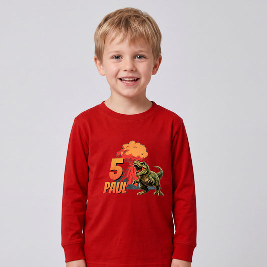 Kindershirt in Rot, einem tiefen klassischen Rot: Ein Junge steht vor hellgrauem Hintergrund, der Vulkankegel verschmilzt mit dem Stoff, während der Tyrannosaurus, die orangefarbene Zahl 5 und der Name PAUL klar lesbar bleiben.