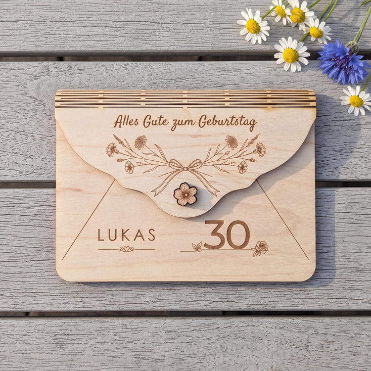 Geschenkverpackung aus Holz zum 30. Geburtstag, Motiv Wiesenblumen: Kornblumen und zarte Gräser um eine Schleife. Name LUKAS und Zahl 30 graviert, Innentasche 152 x 101 mm, auf grauem Gartentisch mit Kamille.