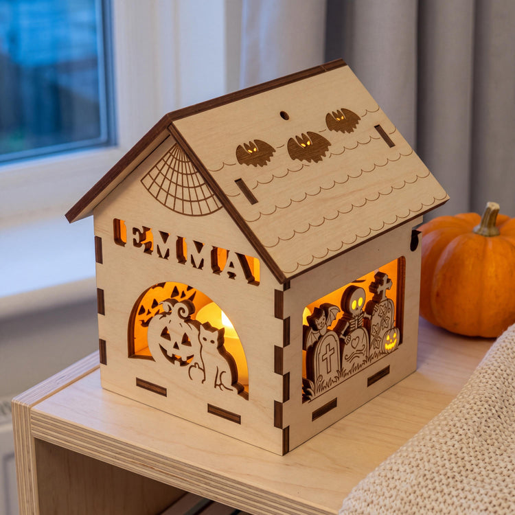 Windlicht mit Namen EMMA auf einem Holzregal im Kinderzimmer am Abend, daneben ein kleiner Kürbis und eine Strickdecke, das Teelicht leuchtet